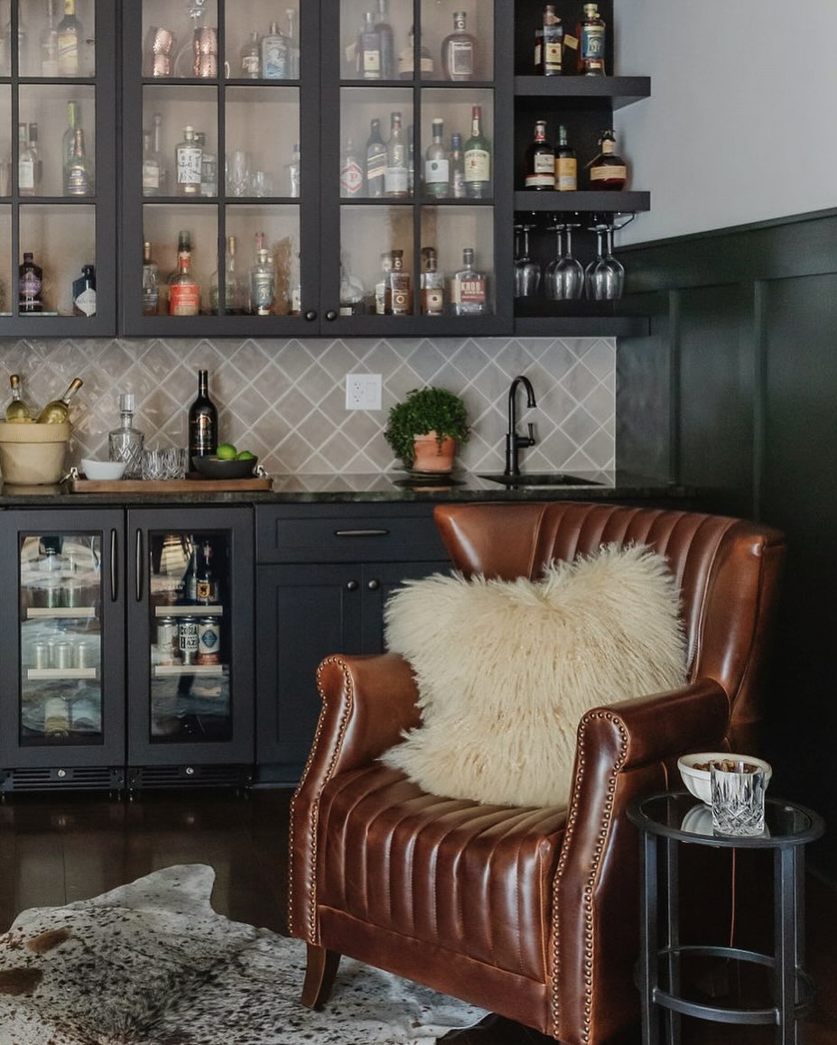 A stylish home bar area features glass-front cabinets of liquor bottles, a countertop with a small sink, and a leather armchair with a fluffy pillow beside a round side table—perfect inspiration for elegant home bar design.