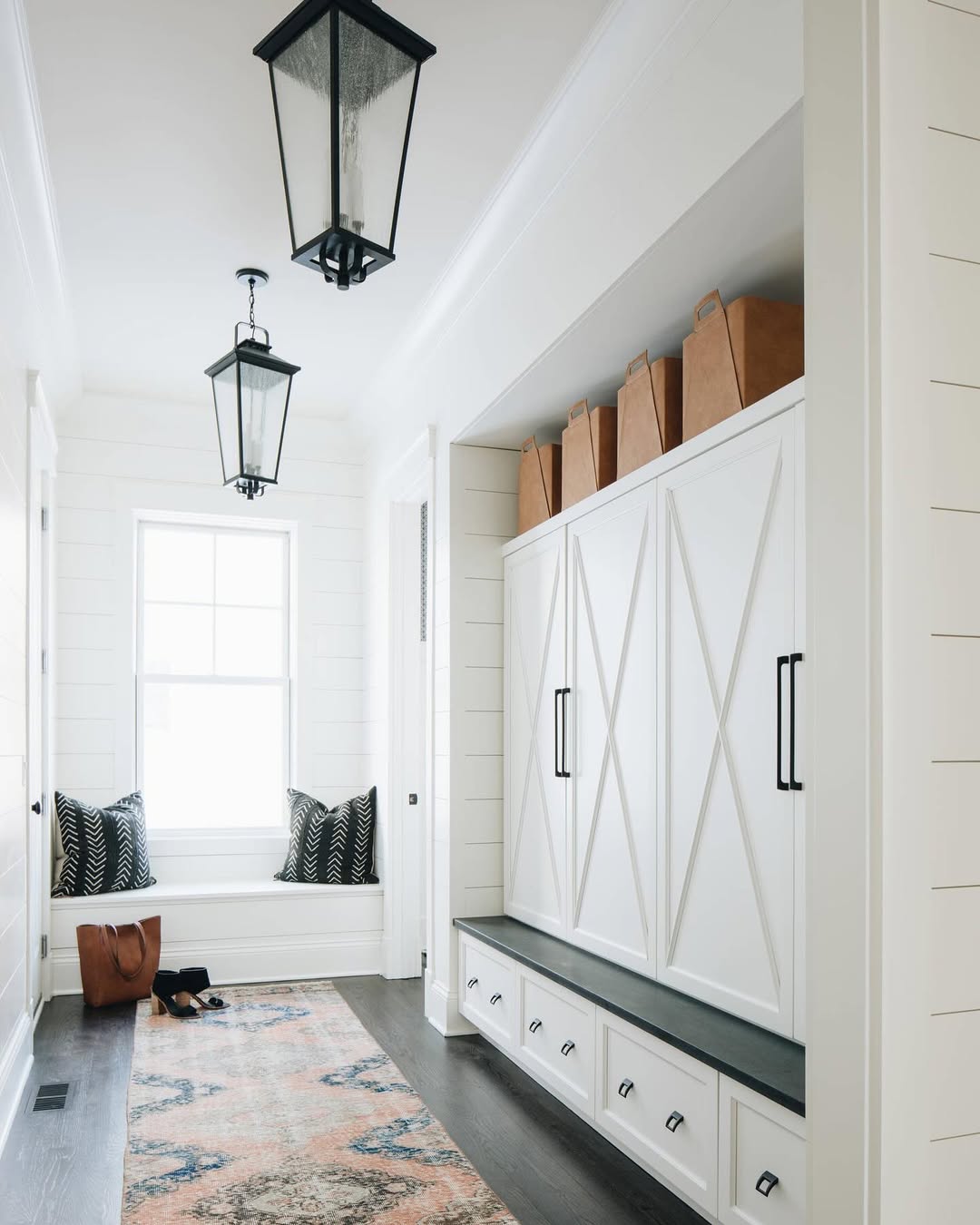 A bright entryway with white built-in cabinets, a window seat with black and white cushions, brown storage bins on top, and a patterned rug on a dark floor—perfect inspiration for genius mudroom ideas.