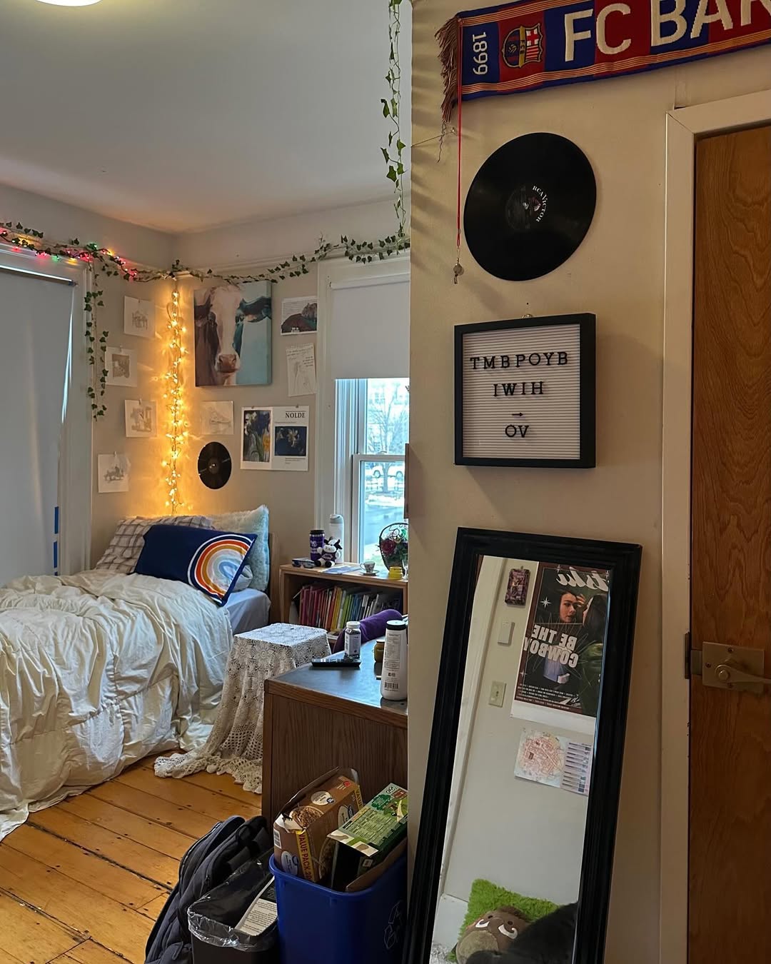 A cozy dorm room for college guys featuring wall art, string lights, vinyl records, plants, a bed with a rainbow pillow, a mirror, and a letter board reading “TMBPOYB I W I H OV.”.