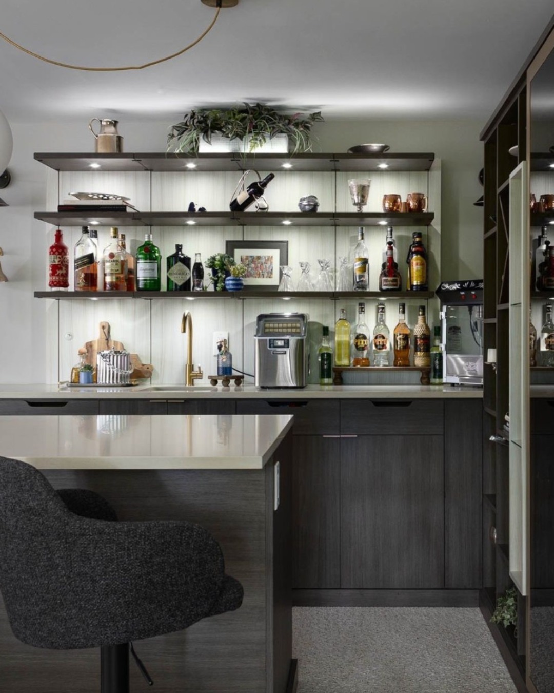 Modern home bar with shelves displaying various liquor bottles, glassware, and decor above a countertop with a coffee machine and sink. A dark upholstered chair sits at the counter—perfect for anyone seeking gorgeous bar ideas for their home.