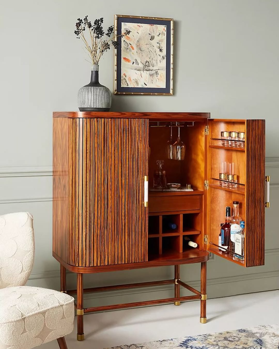 A wooden bar cabinet with a vertical grain pattern stands open, displaying glassware, bottles, and shelves; perfect for home bar ideas, it features a vase and framed art on top as stylish bar decor.