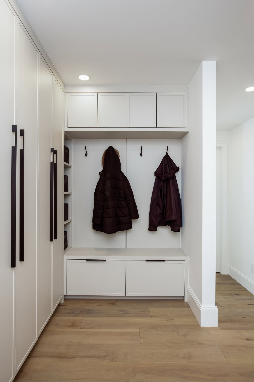A modern mudroom with light cabinetry, two dark jackets hanging on hooks, cubby shelves, and wooden flooring—perfect for inspiring stylish mudroom ideas.