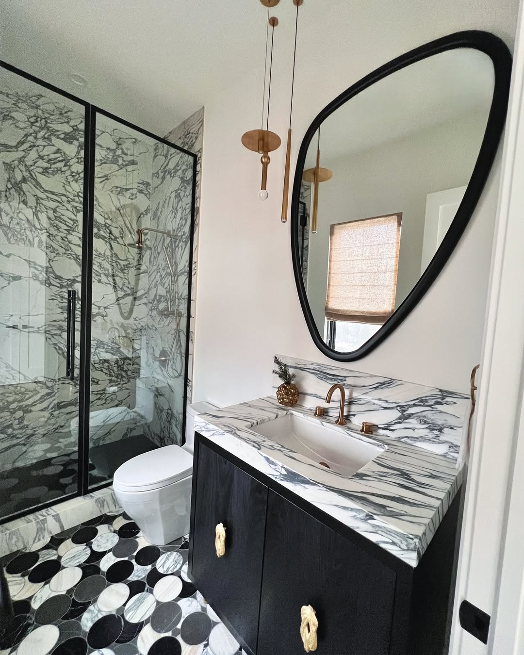 Modern marble bathroom with black and white marble design surfaces, a black-framed glass shower, oval mirror, black vanity with gold hardware, and geometric black-and-white tile floor—perfect for chic bathroom ideas.