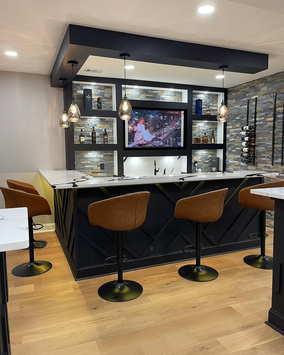 A modern home bar with brown leather barstools, stone accent walls, pendant lights, a mounted TV, and shelves displaying bottles and glasses—perfect inspiration for home bar ideas or reality show-worthy bar decor.