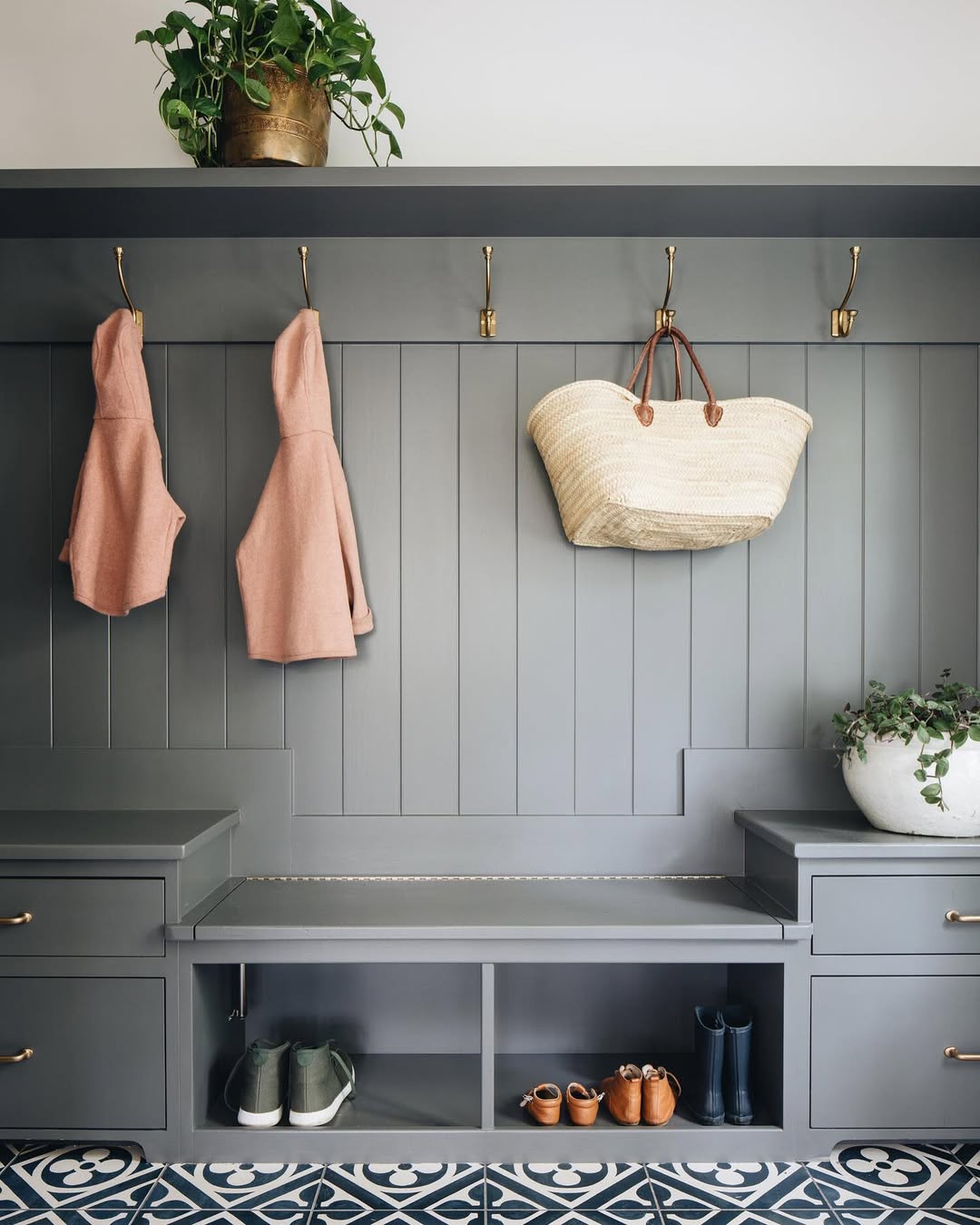 A genius mudroom with gray paneled walls, two pink hooded jackets, a woven bag on hooks, plants, a bench, and shelves for shoes and boots sits atop patterned floor tiles.