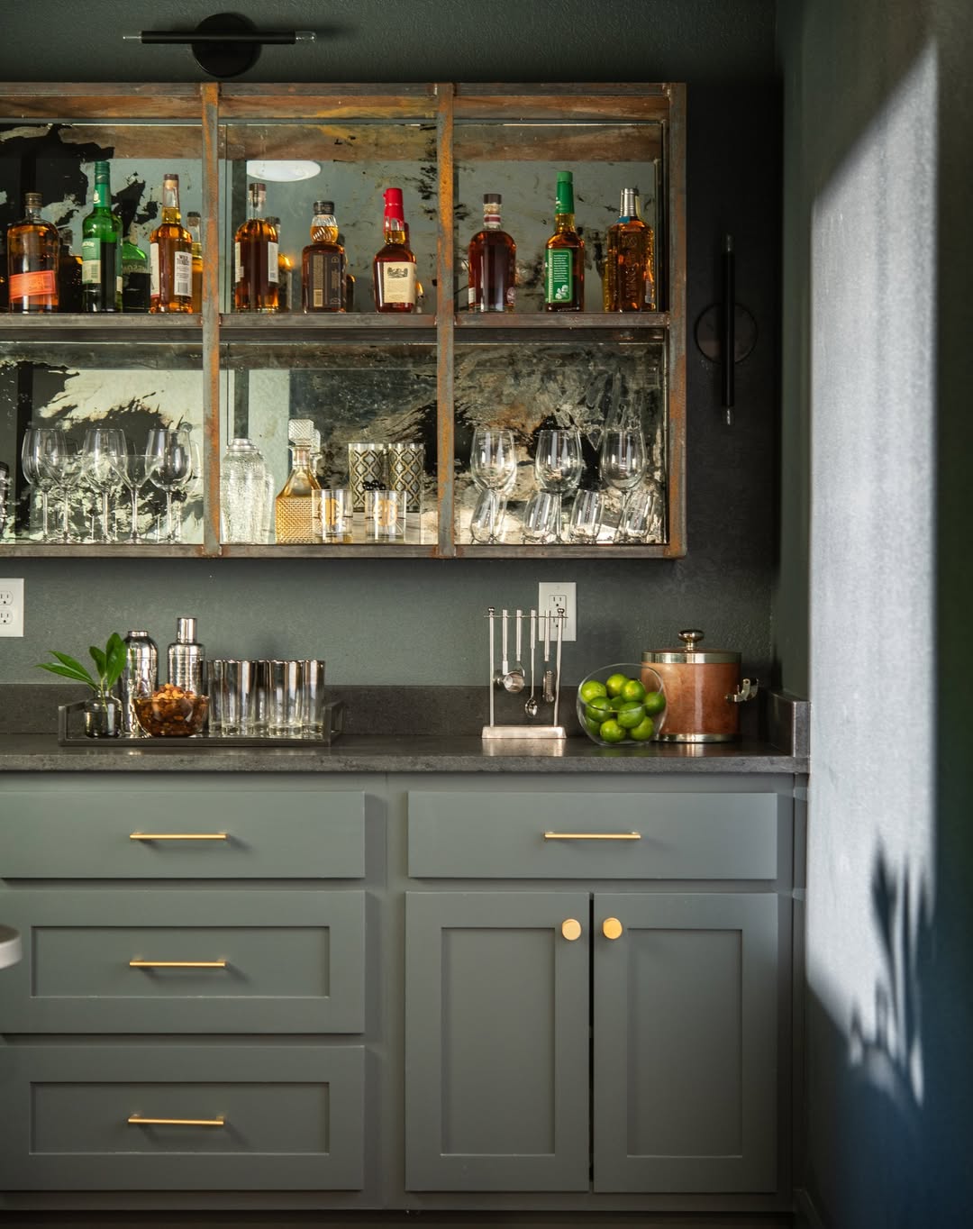 A home bar with bottles of liquor, glassware, cocktail tools, a bowl of limes, and gray cabinets with gold handles—perfect inspiration if you
