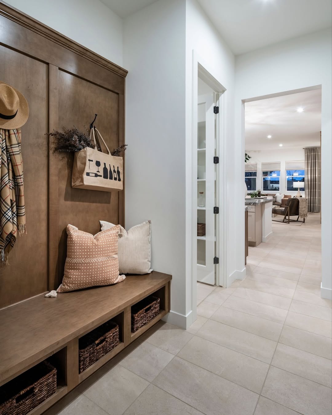 A genius mudroom idea featuring a wooden bench, pillows, woven baskets, hanging scarves, and a tote bag creates the perfect entryway leading to a bright open kitchen and living area.