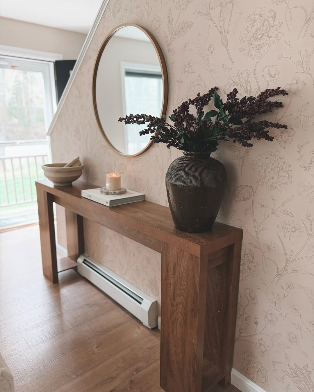 A wooden console table, perfect for stylish console tables inspiration, displays a large vase of dark flowers, a stack of books, a candle, and bowls against floral wallpaper, with a round mirror above.