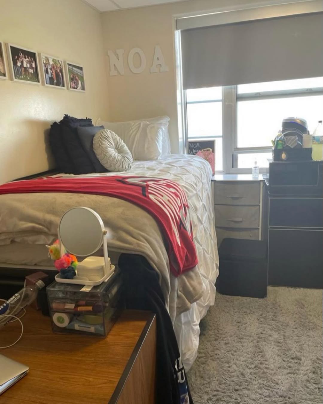 A neatly made dorm room bed with pillows, a blanket, and a red shirt, perfect for a dorm room for college guys, sits next to a window, desk, and drawers, with decor and personal items visible.