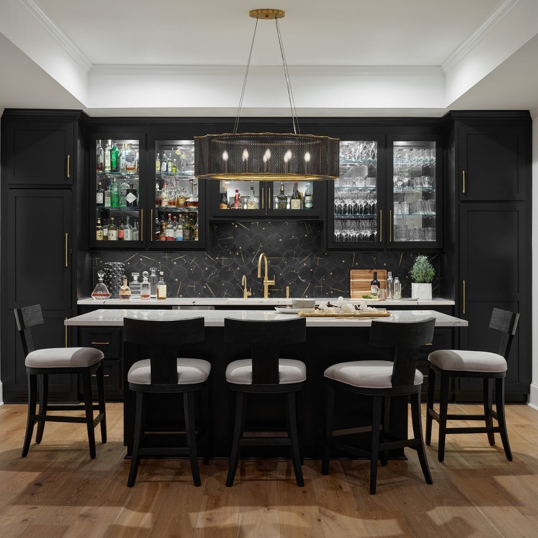 Modern home bar with black cabinets, glass shelves displaying bottles and glassware, a white countertop island with five barstools, and contemporary pendant lighting—perfect inspiration for gorgeous home bars or fresh home bar ideas.