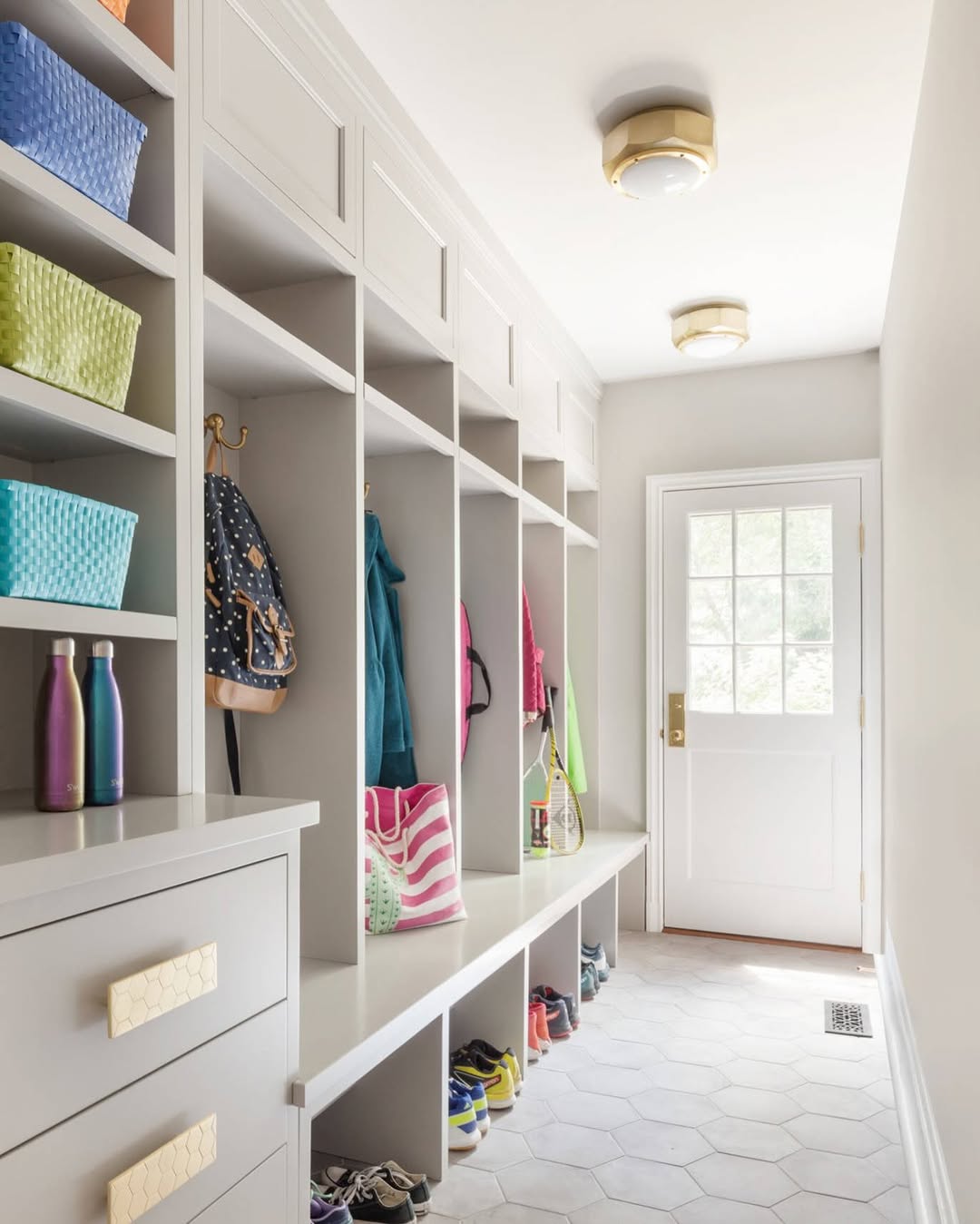 A bright mudroom with cubbies, baskets, water bottles, backpacks, and shoes organized along one wall offers smart entryway storage. A white door and hexagonal tile floor complete this stylish take on practical mudroom ideas.