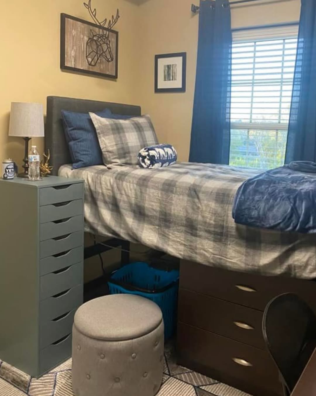 A neatly made bed with plaid bedding is elevated over dressers in this dorm room for college guys, next to a small cabinet, lamp, and stool. A window with dark curtains lets in natural light while wall art decorates the space.