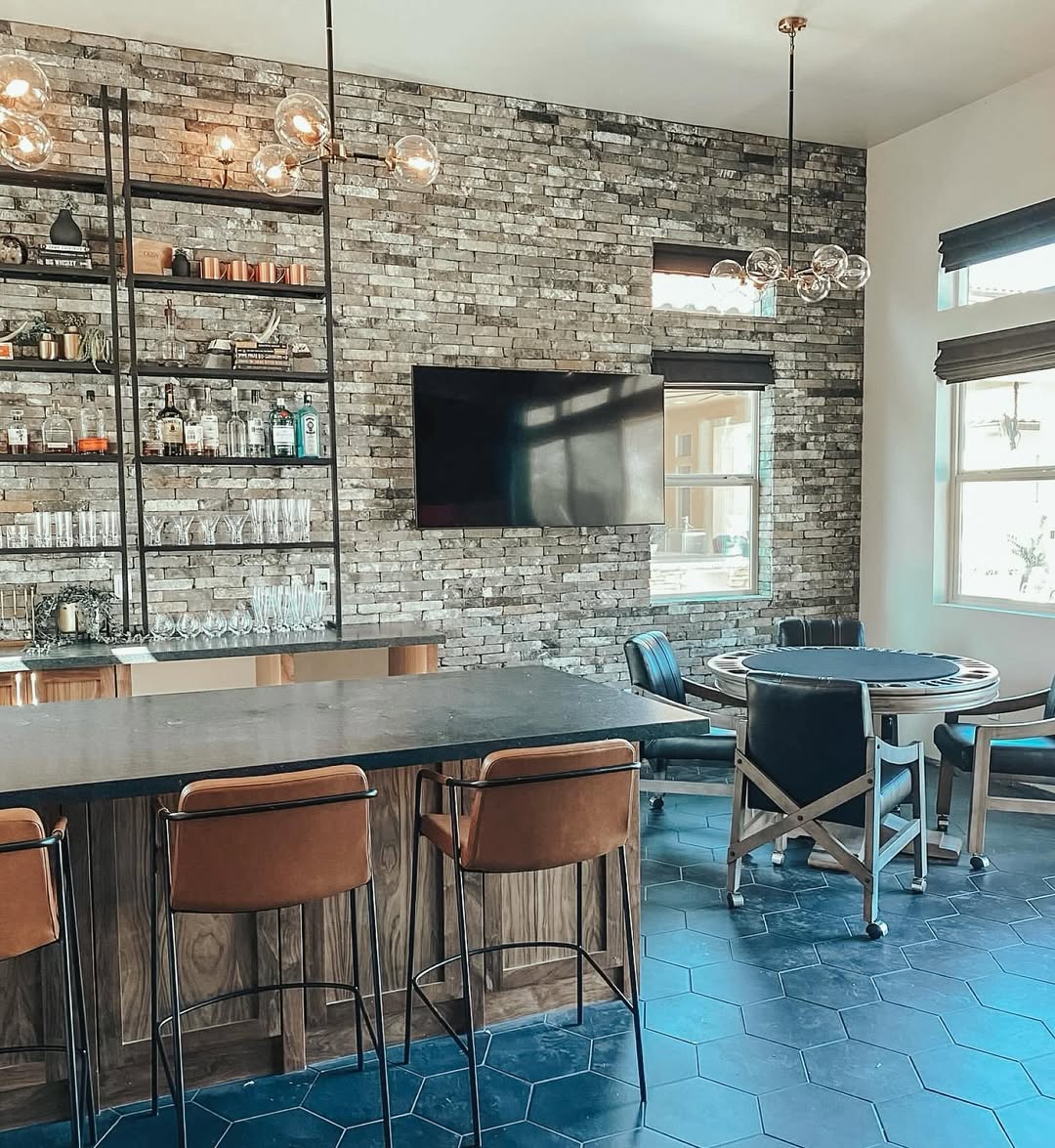 Modern home bar area with brick accent wall, mounted TV, shelves with glassware and bottles, counter with barstools, and a round card table on a hexagon-tiled floor—perfect for those seeking gorgeous bar ideas for home.