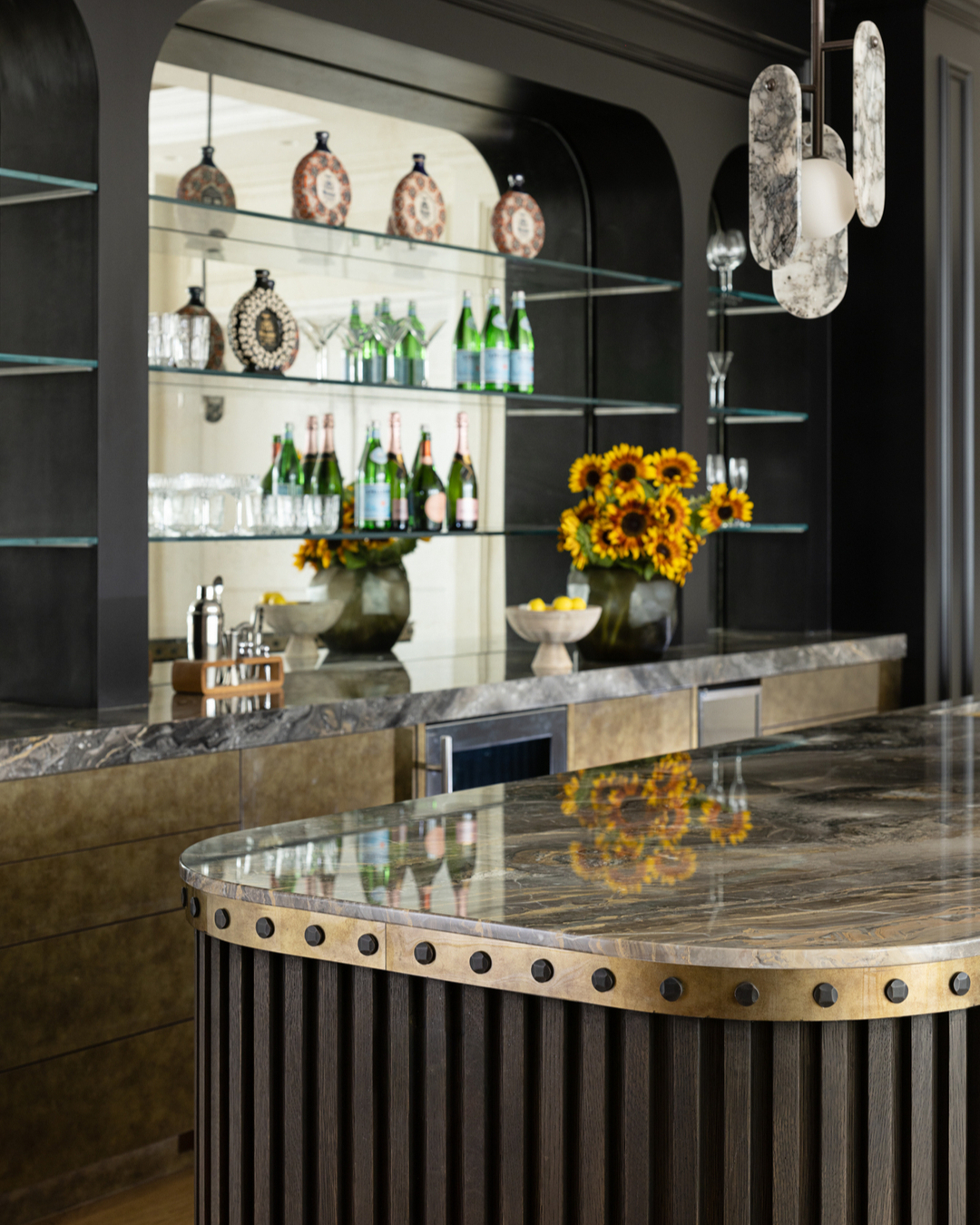 A modern home bar with a marble countertop, glass shelves holding bottles and glasses, decorative plates, sunflowers in vases, and a contemporary pendant light—perfect inspiration for gorgeous bar ideas.