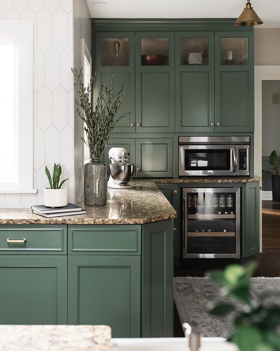 A modern kitchen with green cabinets, granite countertops, stainless steel appliances, potted plants, and glass-front upper cabinets.