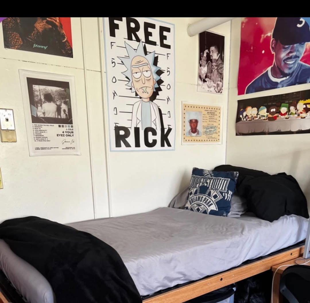 A neatly made dorm bed with gray and black bedding sits in a dorm room for college guys, against a wall decorated with posters and photos, including a large 