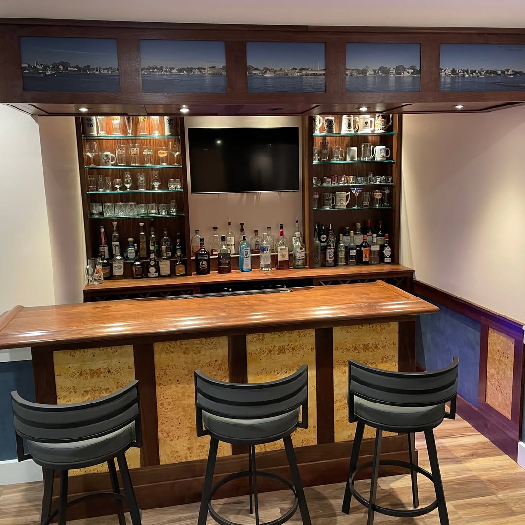 A home bar with a wooden counter, three black barstools, shelves for glassware, an array of liquor bottles, and a mounted TV screen—perfect inspiration for those seeking gorgeous bar ideas for home.