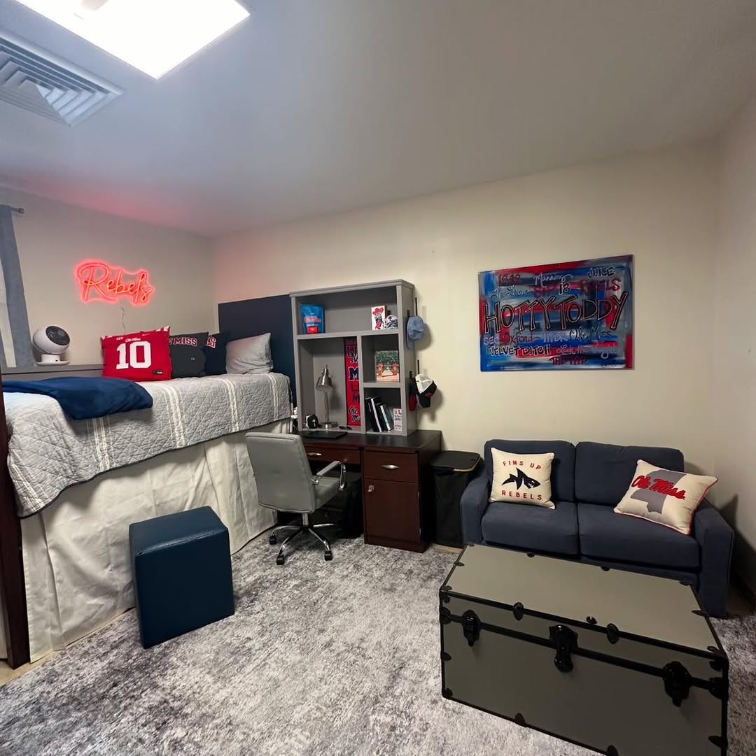 A neatly organized dorm room for college guys with a lofted bed, desk, chair, small couch, trunk, and college-themed decor on the walls and pillows.