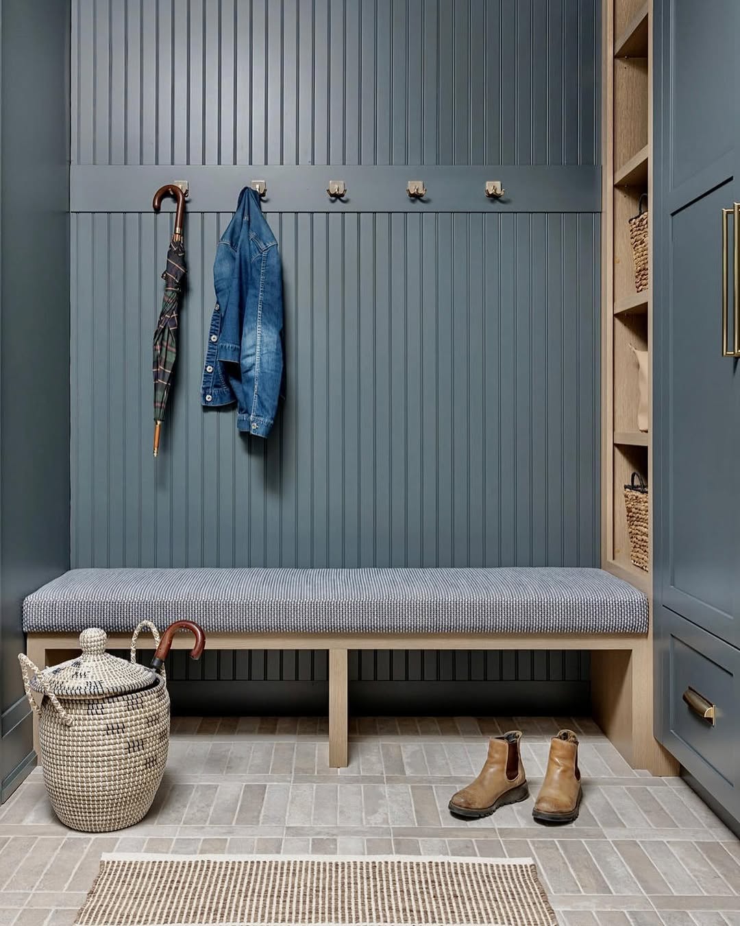 A genius mudroom with blue paneled walls, a cushioned bench, hooks for jackets and umbrellas, woven baskets, a laundry basket, and brown boots on the tiled floor—perfect entryway inspiration for organized living.