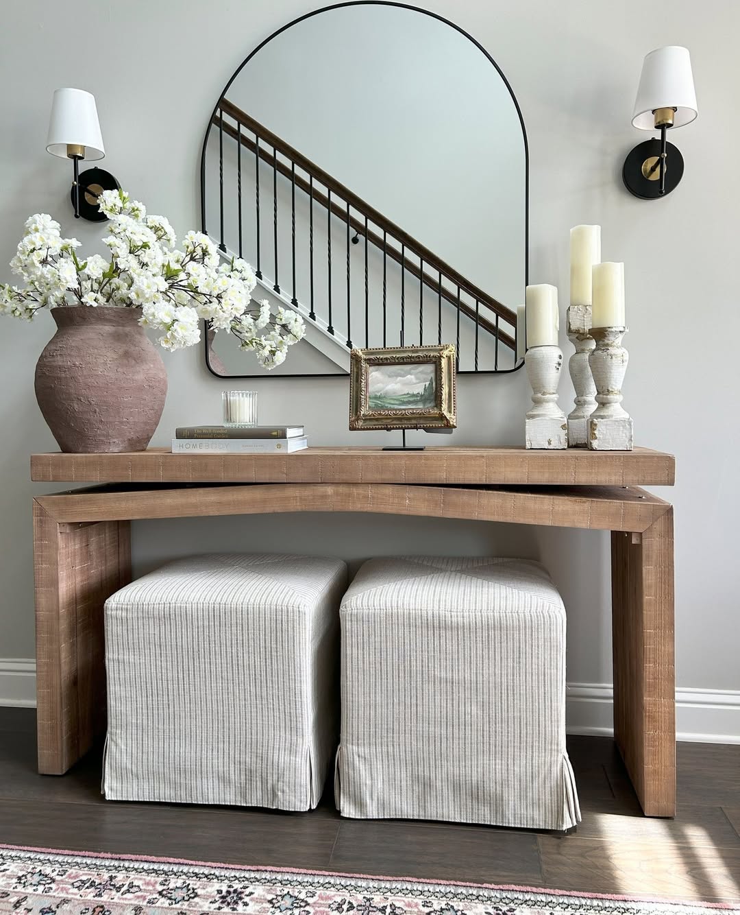 A stylish console table with two upholstered ottomans, a large vase of white flowers, candles, a small painting, books, and a round mirror above, offers inspiring console table decor flanked by elegant wall sconces.