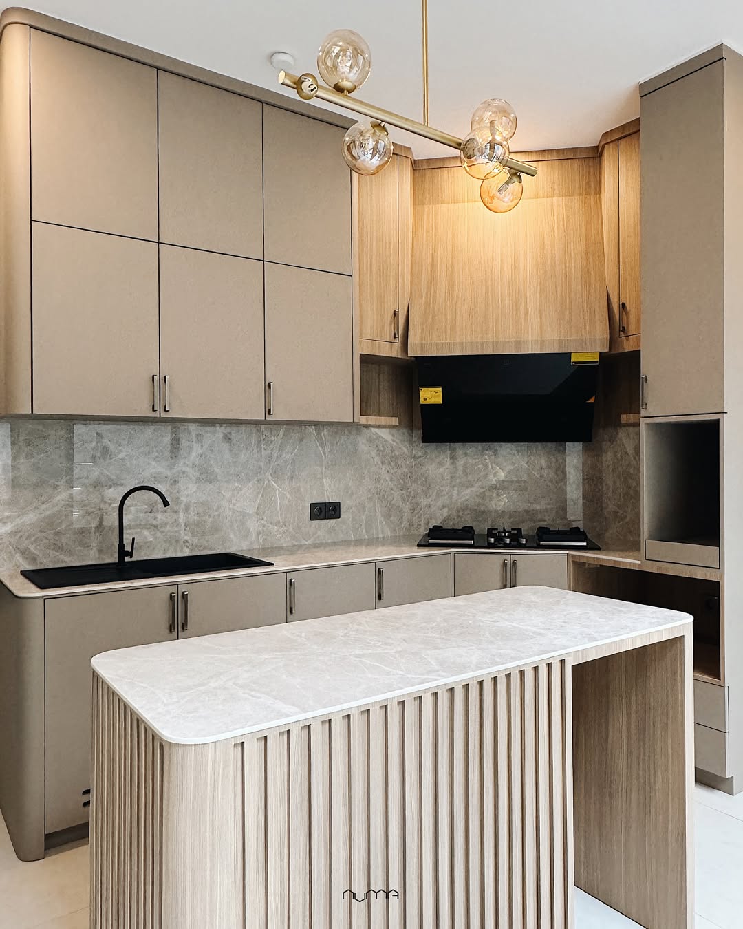Modern kitchen with beige cabinets, marble backsplash and countertop, black sink and stove, built-in oven, and a gold chandelier above a central island.