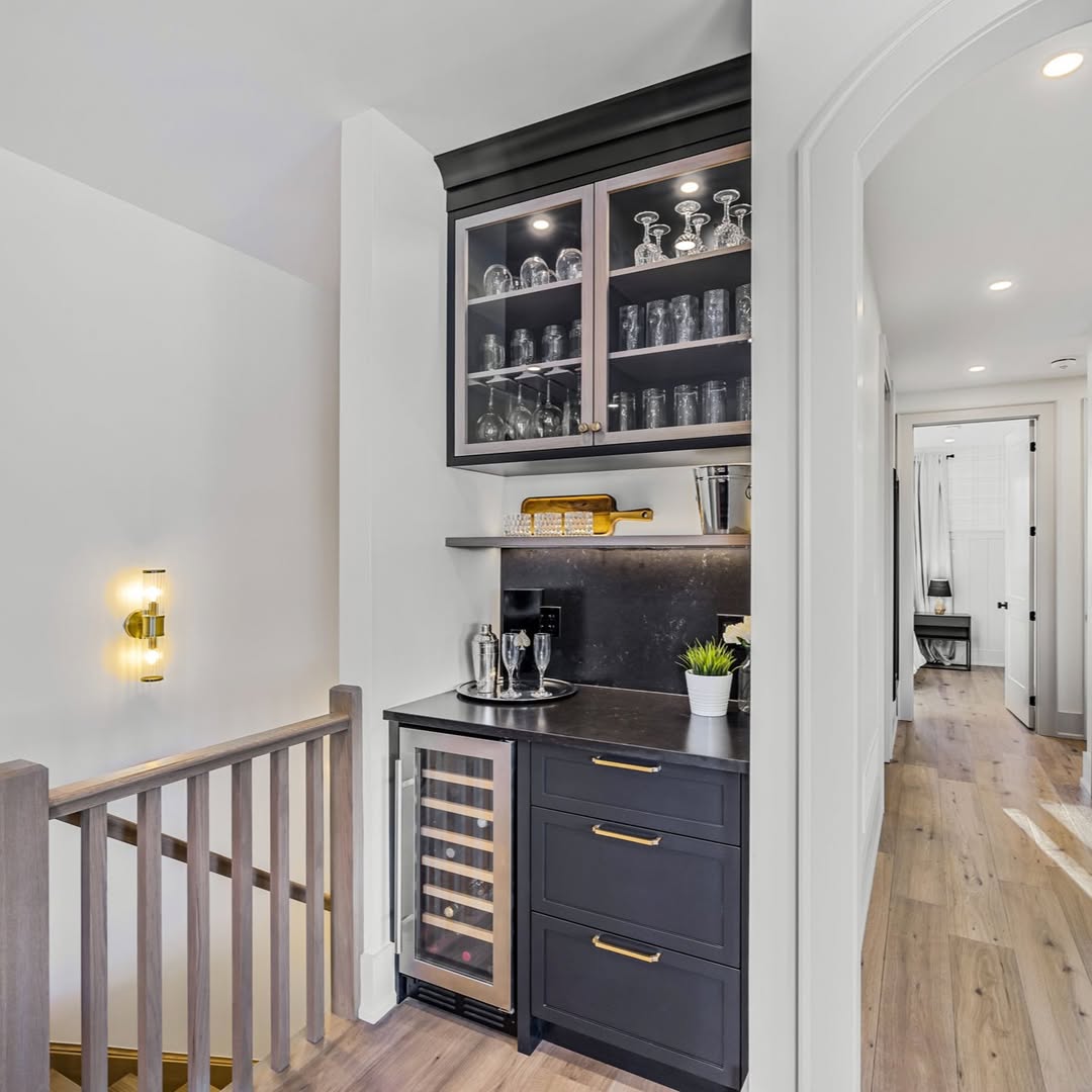 A small home bar with black cabinets, a wine fridge, glassware in upper cabinets, and a countertop with a cocktail shaker, plant, and tray—perfect for those seeking home bar ideas—located by a hallway and staircase.