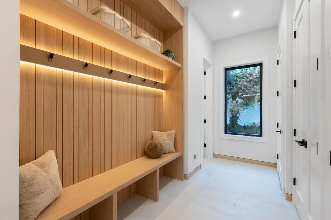 A modern genius mudroom with wood paneling, a built-in bench with pillows, open shelves, wall hooks, white walls, and a large window—a stylish entryway designed for both function and comfort.