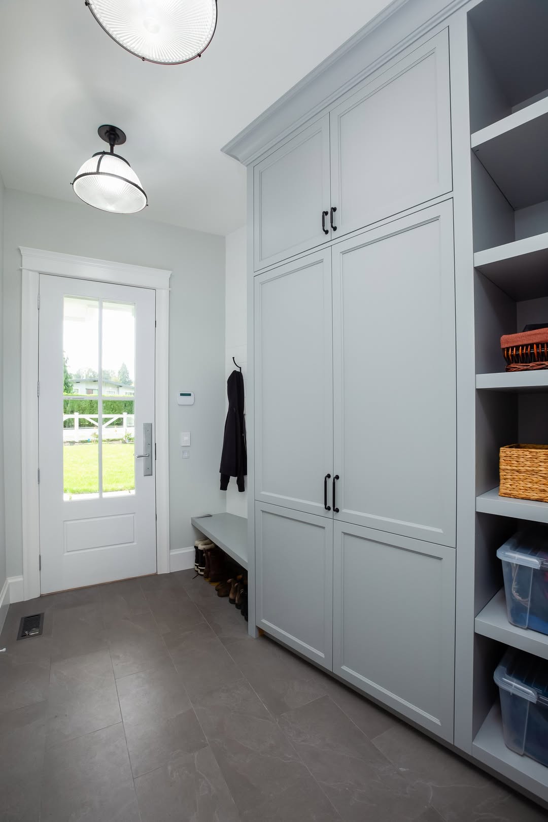 A genius mudroom featuring gray built-in cabinets, open shelves with baskets and bins, a wall hook with a coat, a bench, and an entryway door with a window leading outside.