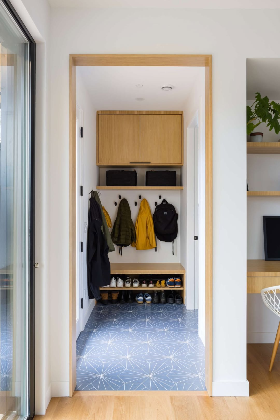 Small entryway mudroom with backpacks and jackets hanging on hooks, shoes on a rack below, two cabinets above, blue geometric tile flooring, and a partial view of a home office on the right—perfect for practical mudroom ideas.