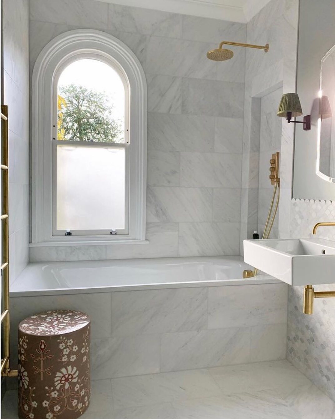 Modern marble bathroom with a marble tub, gold fixtures, arched window, wall-mounted sink, and a decorative floral stool—perfect for those seeking elegant bathroom ideas.