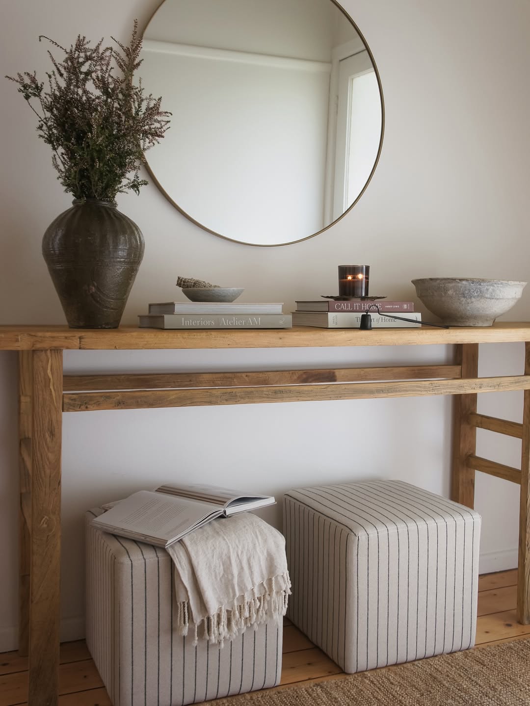 A wooden console table with decorative items, a round mirror above, books, a vase with branches, a bowl, a candle, and two striped ottomans beneath—perfect for anyone seeking stylish console table ideas.
