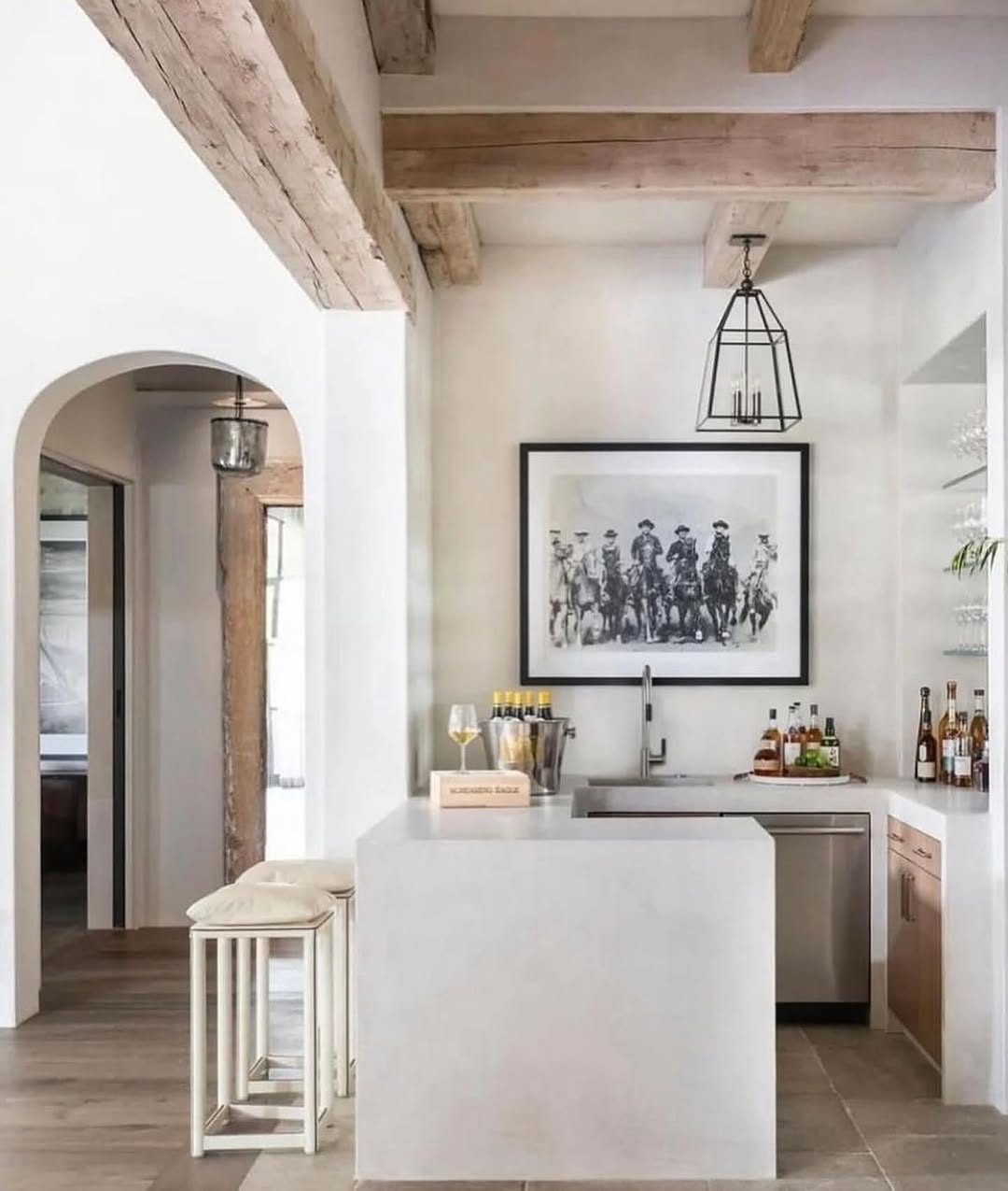 Modern home bar with a white countertop, sink, bottles, wine glasses, and framed black-and-white cowboy artwork above—perfect bar decor inspiration. Exposed wooden beams on the ceiling and light wood floors complete this chic home bar design.