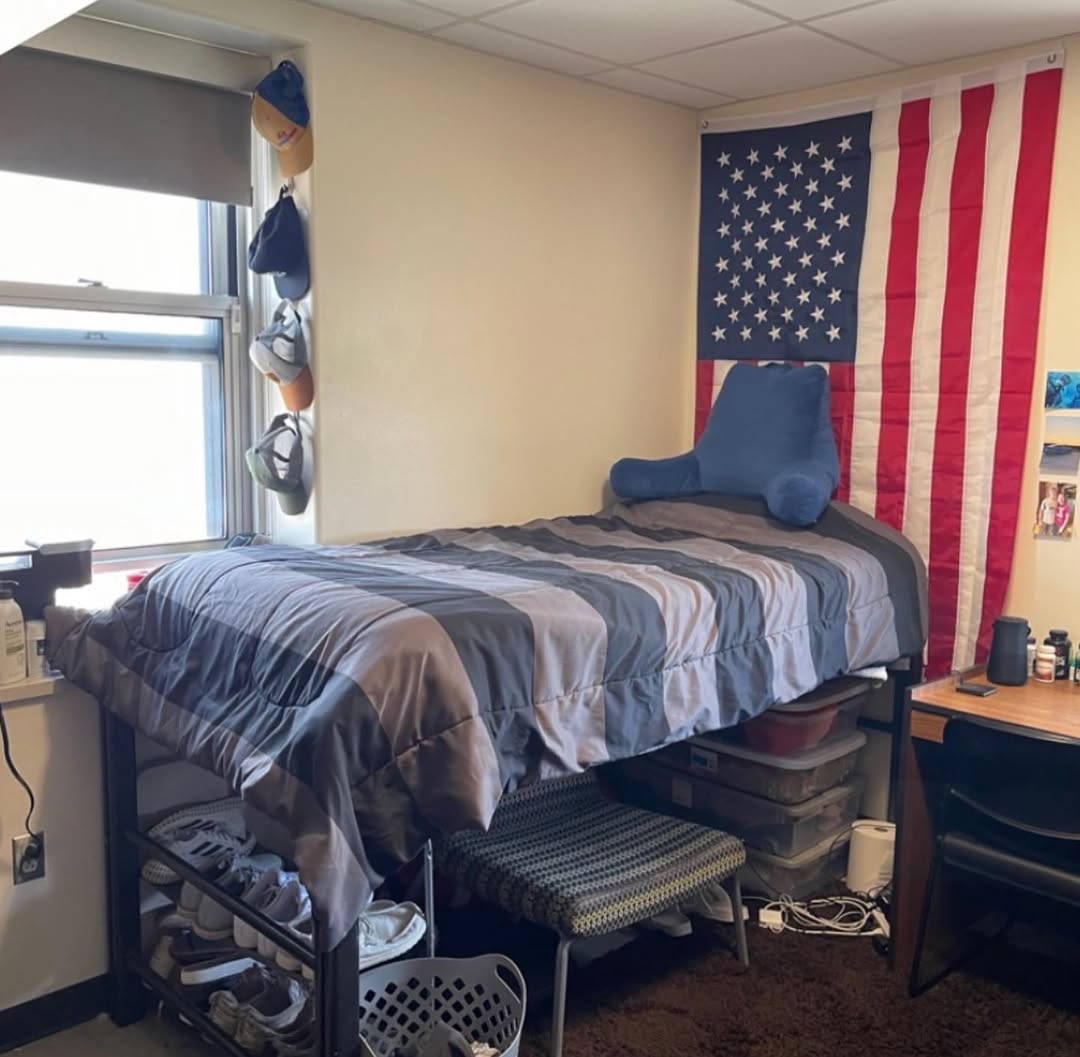 A modern dorm room for college guys with a dark blue and gray color scheme, featuring a bed with storage drawers, a desk, shelves, wall art, and a bulletin board with notes.