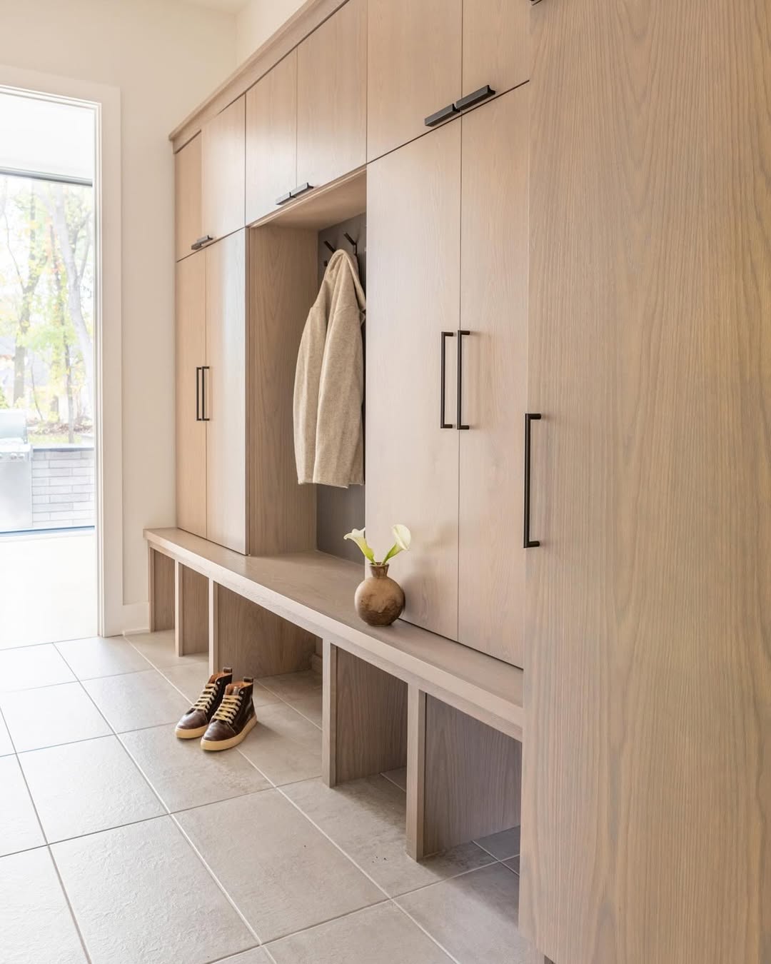 Light wood mudroom lockers with black handles, a bench, a beige jacket, a vase with flowers, and sneakers on tiled floor—perfect entryway inspiration for stylish mudroom ideas.