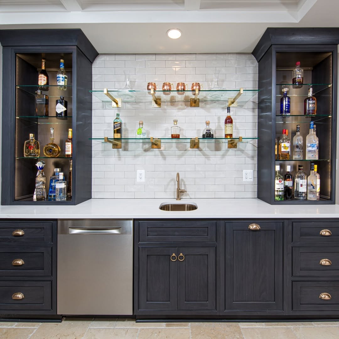 Modern home bar with dark cabinets, a small sink, stainless steel dishwasher, and shelves displaying liquor bottles against a white subway tile backsplash—perfect for those seeking gorgeous bar ideas for home.