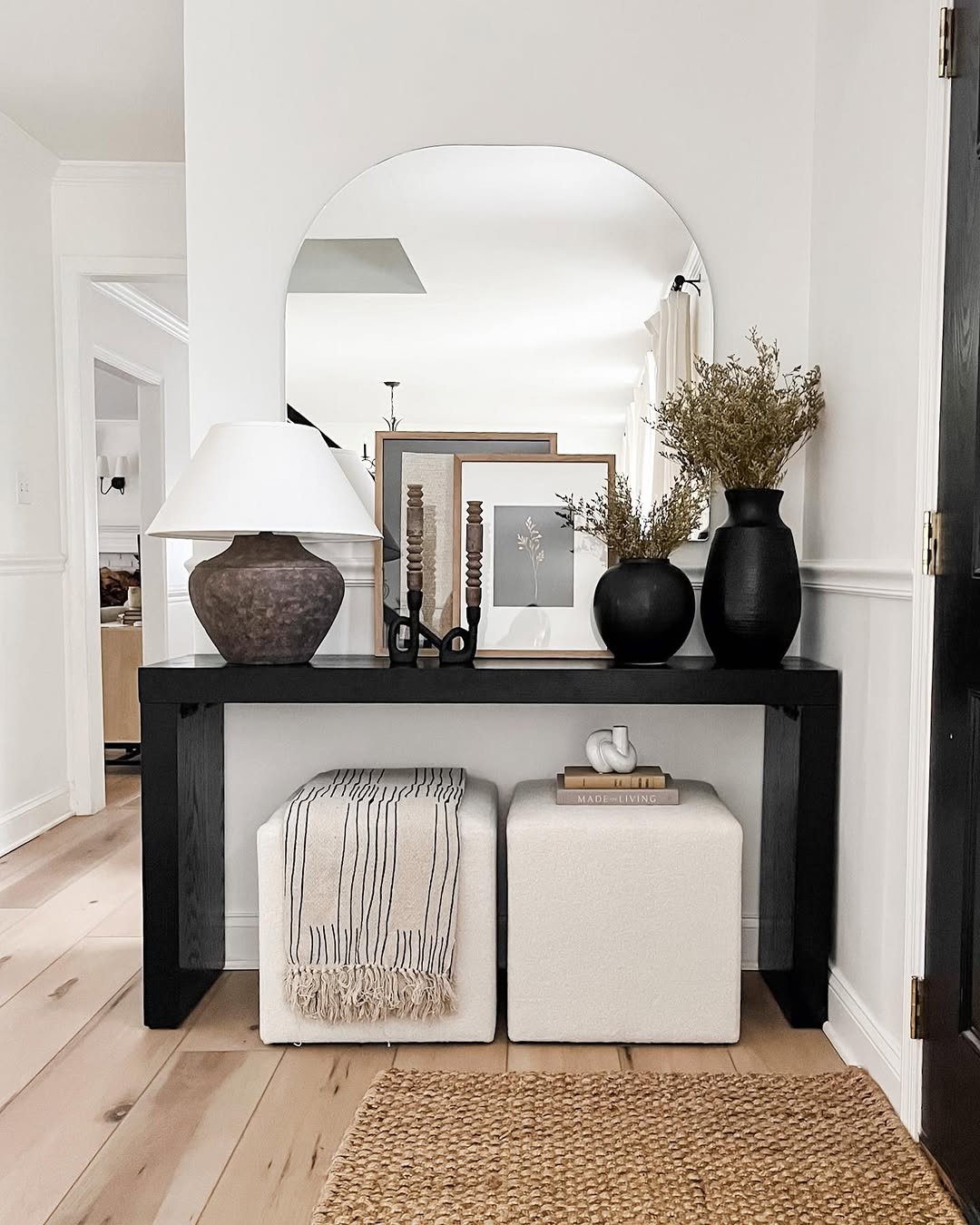 A black console entry table decor setup features vases, a lamp, candles, and framed photos. Two cushioned stools and stacked books rest underneath, while a large mirror completes the elegant display behind.