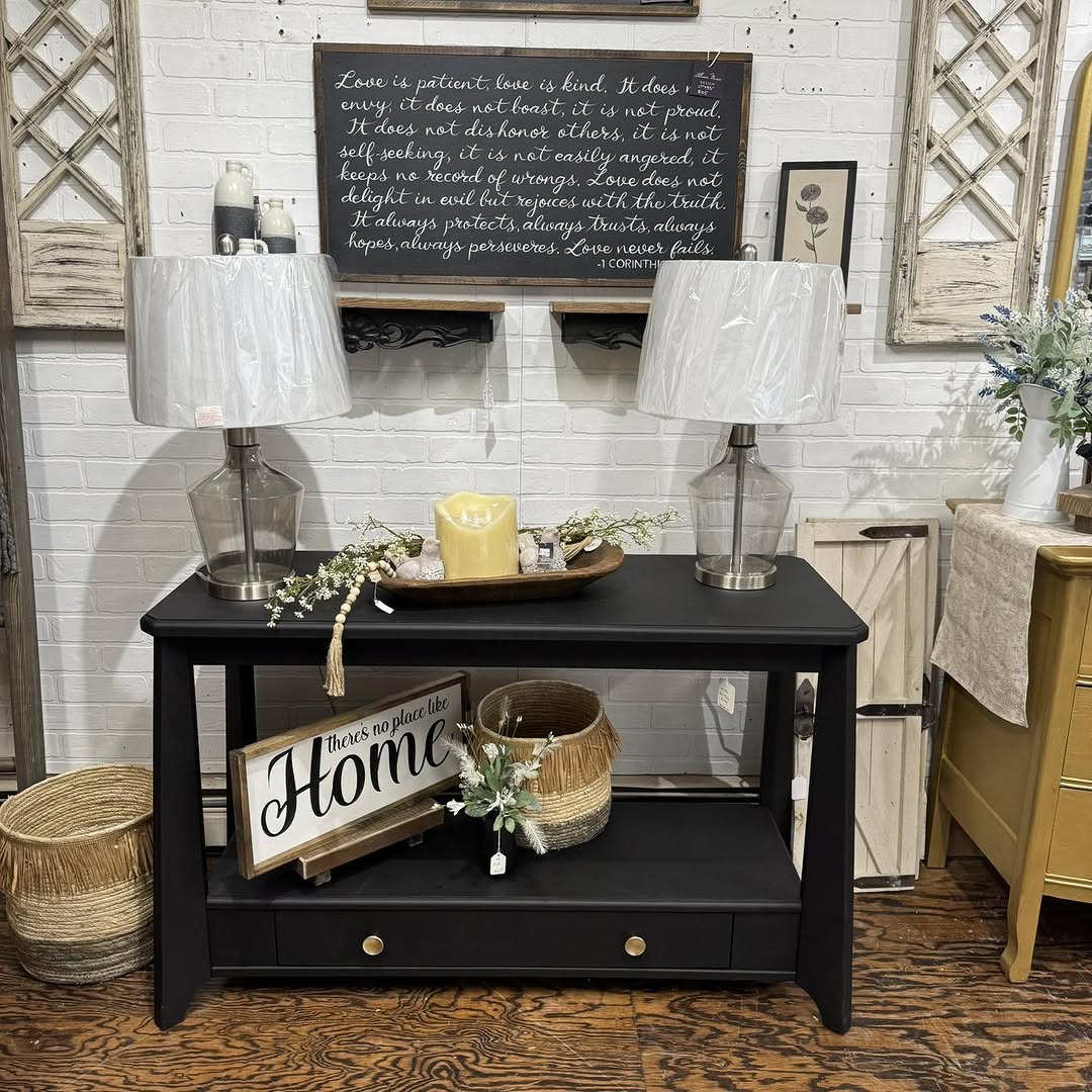 A wooden entry table with decor like a lamp, books, and vases sits below a large round mirror, reflecting a dining area with a table and plants.