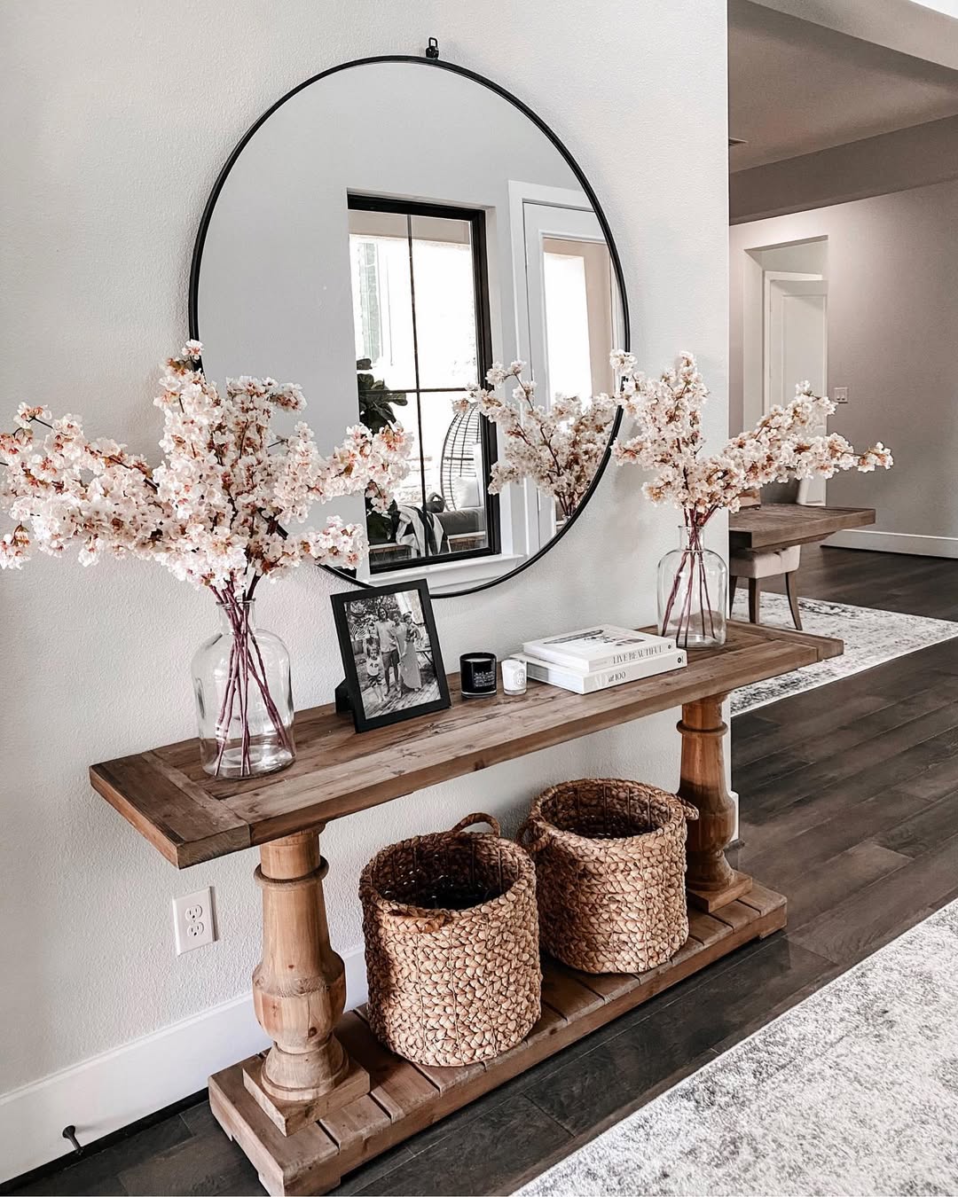 A wooden console table with entry table decor like two baskets, floral arrangements, a framed photo, books, and a round mirror hanging above it in a modern entryway.