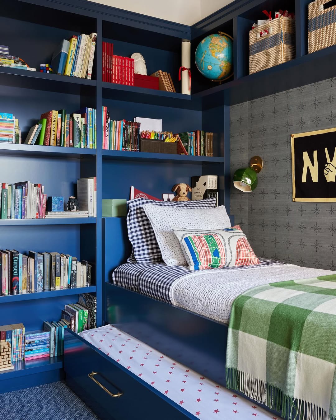 A navy blue built-in bookshelf surrounds a teen boy bedroom, featuring patterned bedding, a green checkered blanket, wall-mounted lamp, and decorative items like a globe and woven baskets.