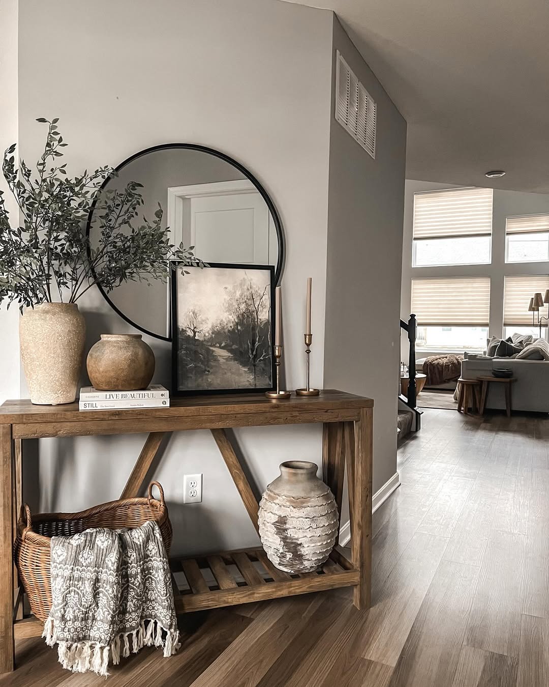 A wooden entry table adorned with vases, a round mirror, a framed picture, books, and a basket with a blanket stands in a modern, neutral-toned living space with large windows—perfect for stylish entry table decor.