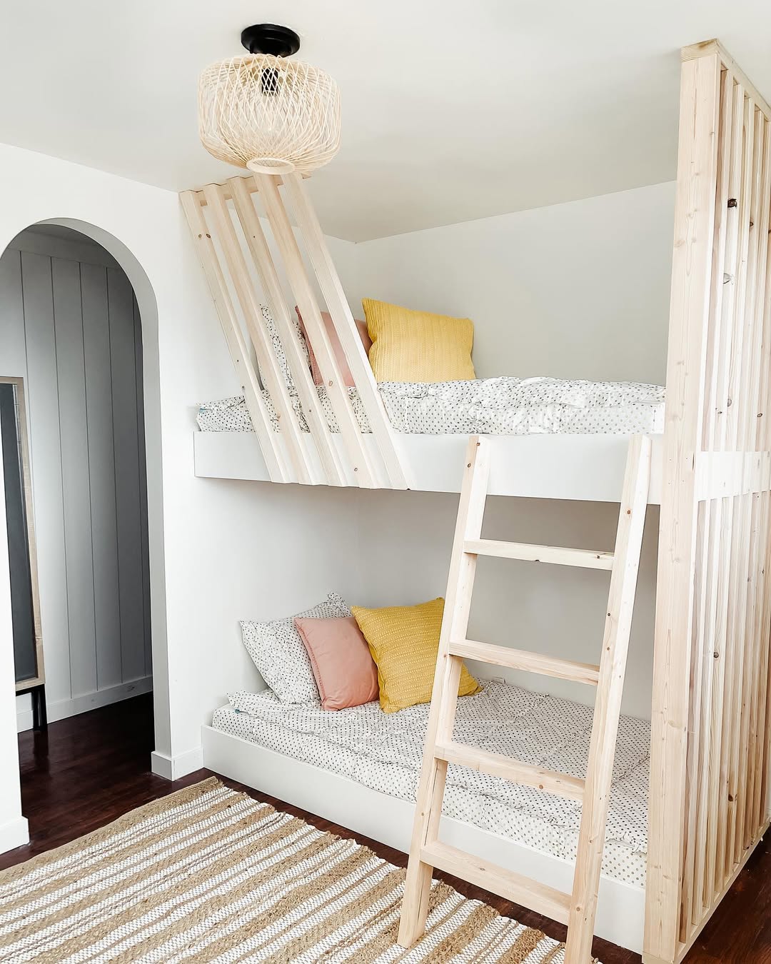 A modern bedroom featuring a light wood loft bed for small rooms, patterned bedding, yellow and pink accent pillows, a striped rug, and a woven ceiling light fixture.