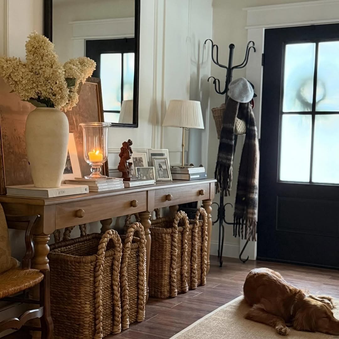 A wooden console table with baskets underneath sits in an entryway, styled with entry table decor like a vase of flowers, framed photos, a lit candle, and a dog lying on the floor nearby.