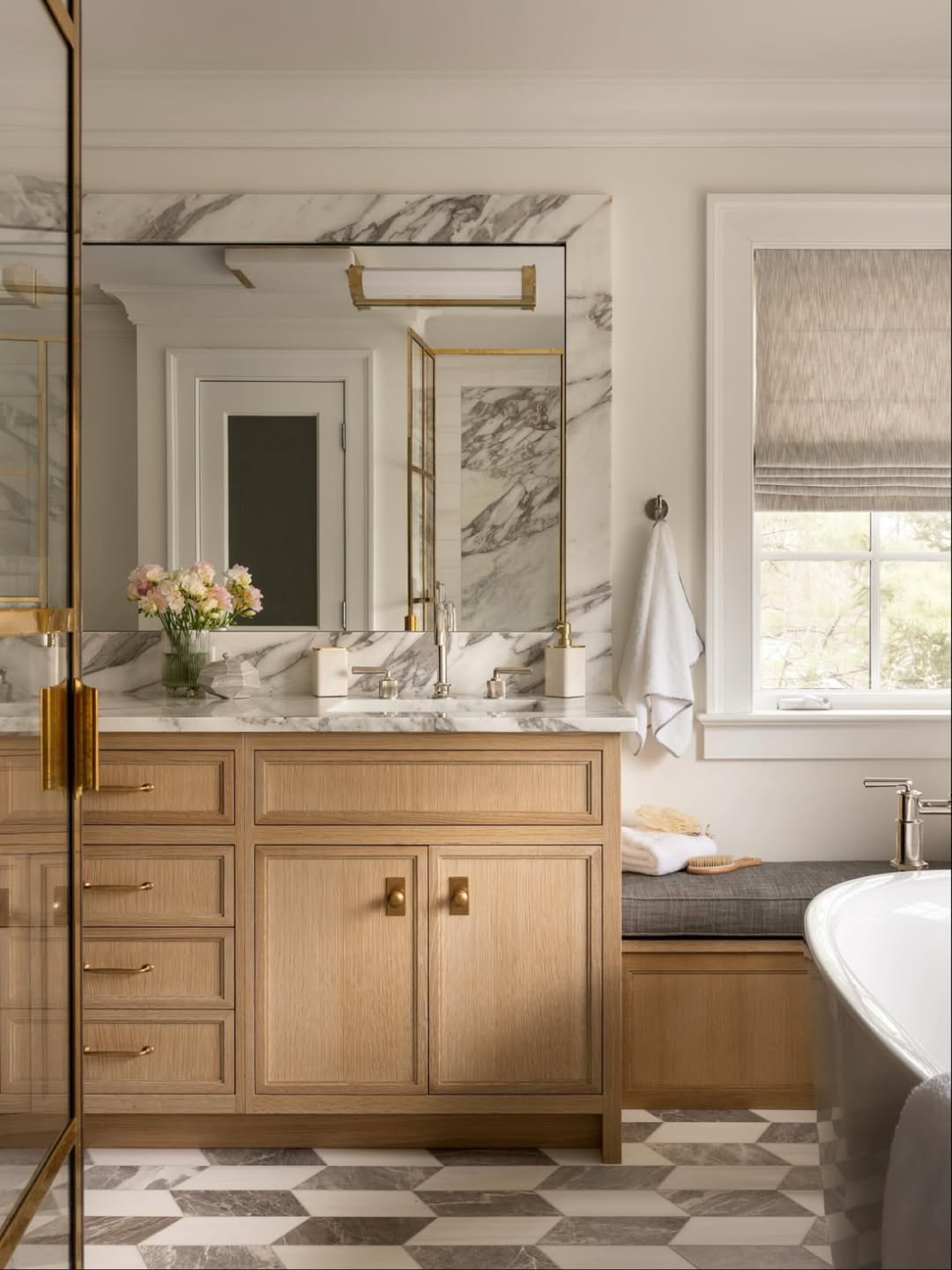 This marble bathroom features a wood vanity, marble countertop, large mirror, gold fixtures, geometric tile floor, and a window with a shade. For more marble bathroom ideas, a vase of flowers adds a fresh touch to the counter.