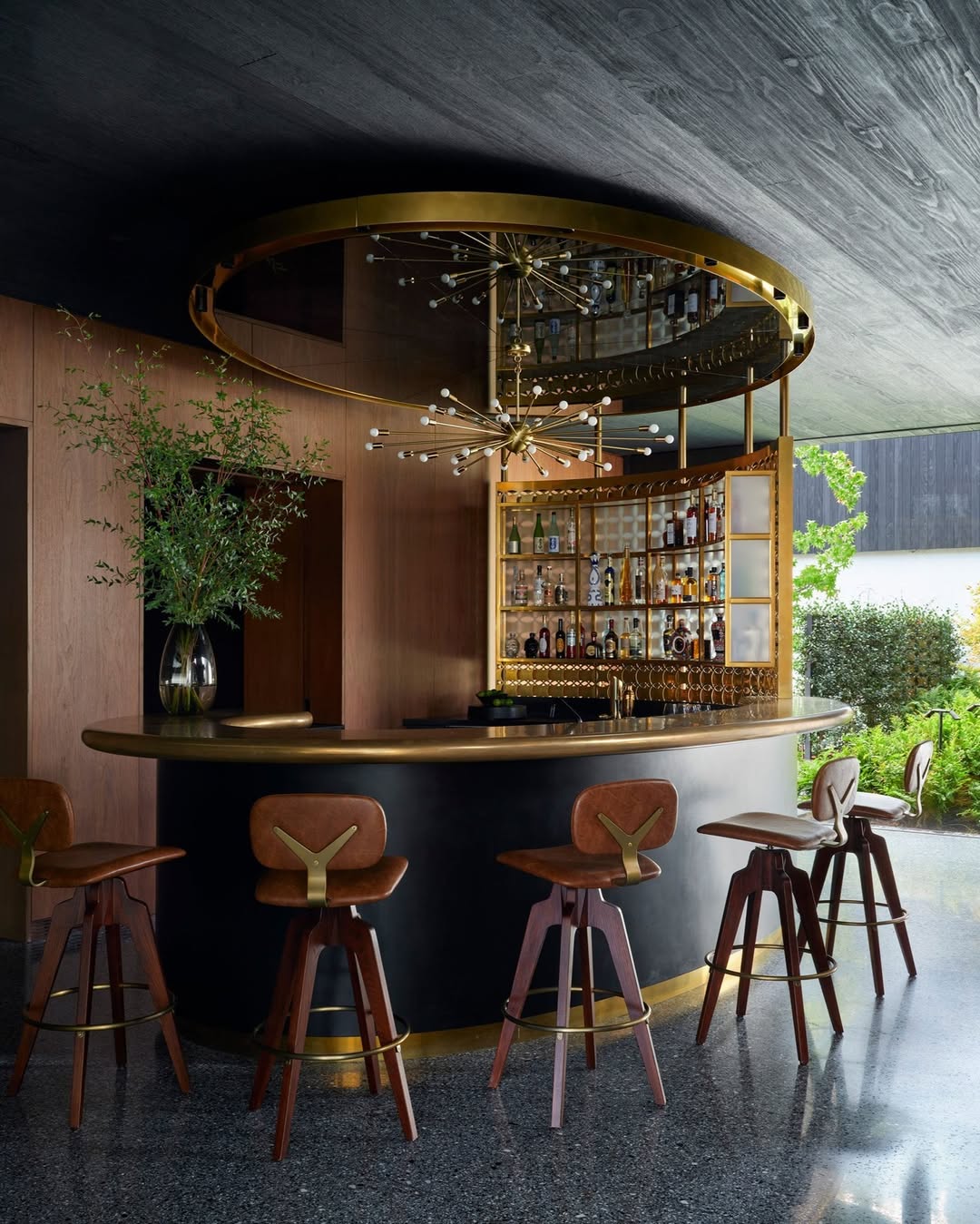 A modern home bar with a curved black counter, gold accents, leather bar stools, mirrored shelves with bottles, and a chandelier overhead. Large windows showcase greenery—perfect inspiration for anyone seeking gorgeous bar ideas.