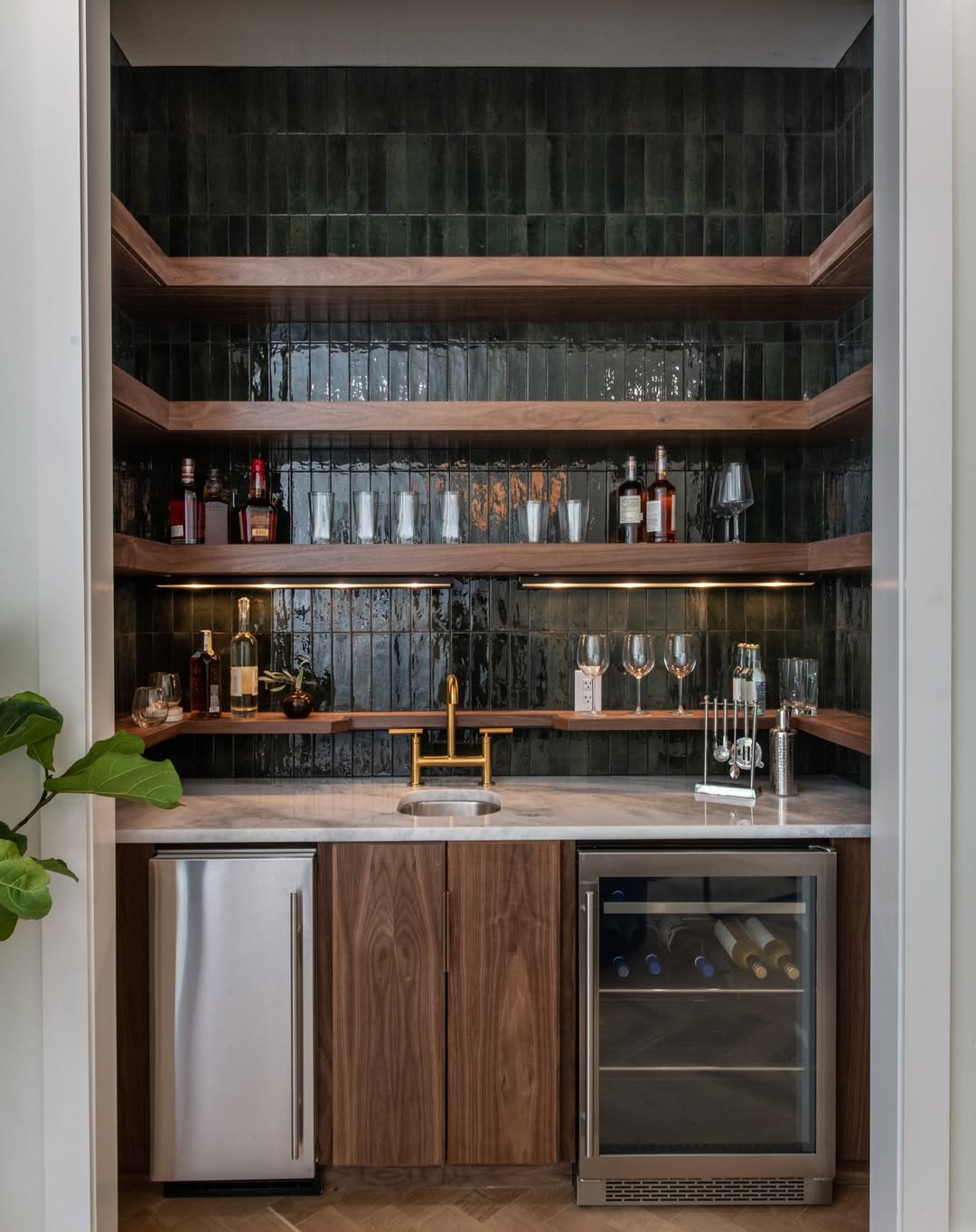 A modern home bar with dark green tile backsplash, wooden shelves for bottles and glasses, a built-in sink, mini fridge, and wine cooler—perfect inspiration if you’re looking for stylish home bar ideas.