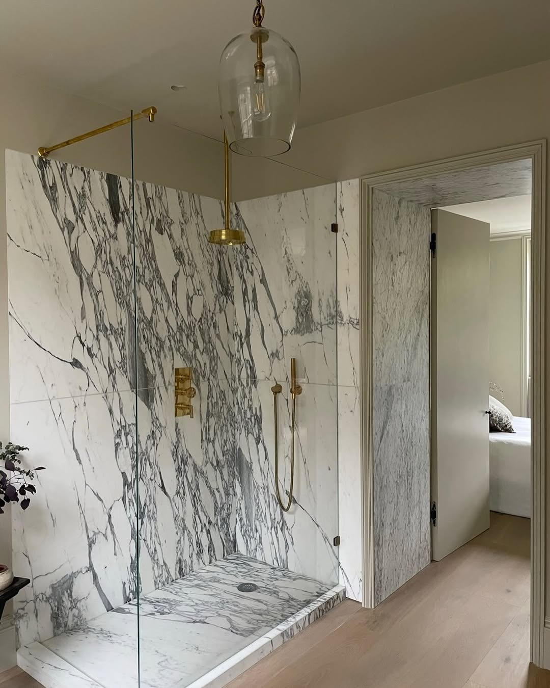 A modern marble bathroom with a walk-in shower featuring white marble walls with dark veining, gold fixtures, a glass partition, and light wood flooring—perfect for those seeking elegant marble bathroom ideas.