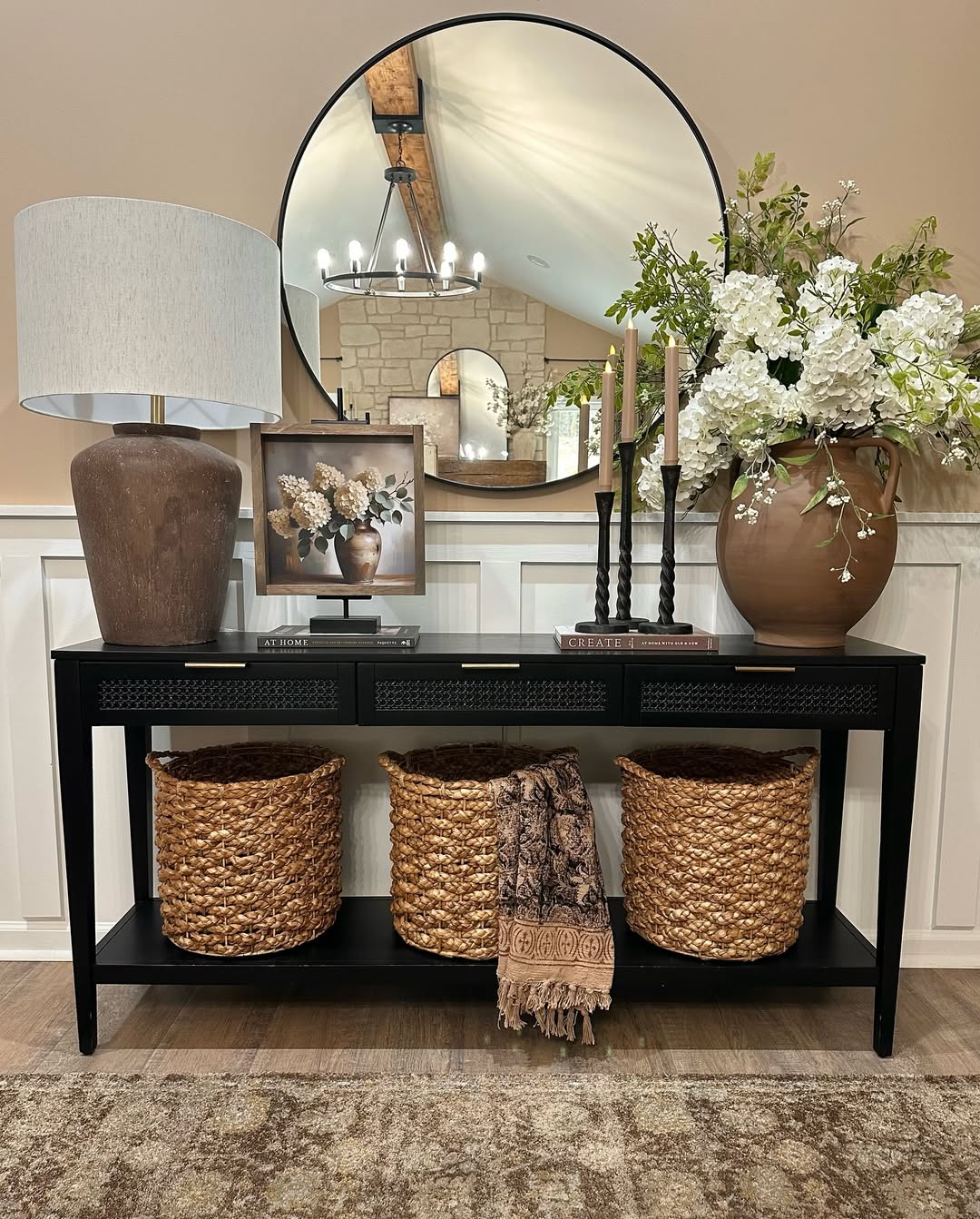 A black console table with three woven baskets, a lamp, framed art, candles, a vase with white flowers, and a round mirror reflecting a chandelier—perfect inspiration for stylish console table decor.